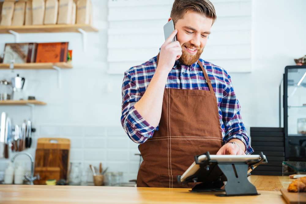 Handsome smiling barista with beard taking order on cell phone and using tablet in cafeteria
